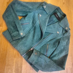 Grön mockajacka från Vero Moda - Snygg grön mockajacka från Vero Moda med klassisk bikerstil. Jackan har silvriga dragkedjor, bred krage med tryckknappar och flera fickor framtill. Materialet är mjuk mocka och jackan är helfodrad för extra komfort.