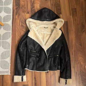 Svart skinnjacka med foder - Snygg svart skinnjacka med beige foder och huva. Jackan har dragkedja och två fickor med dragkedjor framtill. Den har långa ärmar med uppvikta muddar som visar det mjuka fodret. Perfekt för kyligare dagar!