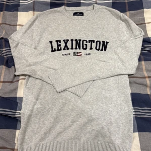 Grå Lexington stickad tröja - Snygg grå stickad tröja från Lexington med svart broderad logga och amerikansk flaggdetalj på bröstet. Rund hals och ribbade muddar vid ärmslut och nederkant. Perfekt för dig som gillar klassisk och clean stil.
