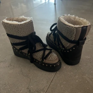 Gråa Inuikii boots med fårskinn - Säljer ett par gråa Inuikii boots med svart snörning och dekorativa nitar runt sulan. Skorna är fodrade med mjukt fårskinn och har en robust svart sula. Perfekta för kalla dagar och ger en cool vinterlook.