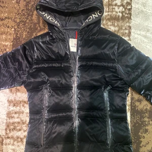 Svart Moncler pufferjacka med huva - Svart pufferjacka från Moncler med stor huva och logotyp på insidan. Jackan har två dragkedjeförsedda fickor framtill. Insidan har en serietidningsetikett, perfekt för kalla dagar och riktigt snygg! Kom privat för fler bilder 