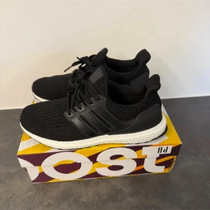 Adidas Ultraboost svarta sneakers - Svarta Adidas Ultraboost sneakers med stickad ovandel och klassiska tre ränder i plast på sidorna. Skorna har vit Boost-mellansula och svart yttersula med greppvänligt mönster. Perfekta för dig som gillar stilrena och bekväma sneakers med modern design.