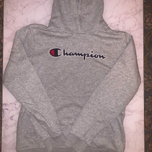 Grå Champion hoodie X-Large - Säljer en grå hoodie från Champion i storlek X-Large. Tröjan har klassisk huva, ribbade muddar och stor Champion-logga broderad på bröstet samt liten logga på ärmen. Perfekt för chill eller streetwear. Materialet är mjuk bomullsmix.