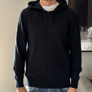 Svart hoodie med huva och snörning - Svart hoodie med klassisk huva och snörning, perfekt för chill dagar. Tröjan har långärmade ribbade muddar och en enkel, stilren look. Passformen är normal och materialet känns mjukt mot huden. Snygg att matcha med jeans eller joggers.