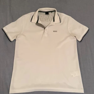 Vit kortärmad piké från BOSS - Vit pikétröja från BOSS med klassisk krage och knäppning framtill. Diskret BOSS-logga på bröstet och svarta detaljer på krage och ärmslut. Perfekt för en clean och stilren look. Pris kan diskuteras