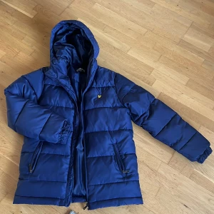 Blå pufferjacka från Lyle & Scott - Blå pufferjacka från Lyle & Scott med huva och dragkedja. Jackan har två fickor med dragkedja och ikonisk gul logga på bröstet. Perfekt för kalla dagar och har en stilren, sportig look. Tillverkad i syntetmaterial för extra värme.