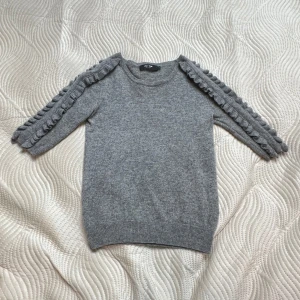 Grå kashmirtröja med volang från Lisa Yang - Superskön grå tröja i 100% kashmir från Lisa Yang. Tröjan har trekvartsärm och volangdetaljer längs axlar och ärmar. Ribbad kant vid ärmslut och nederkant.