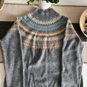 Grå stickad tröja med mönster från ONLY - Mysig grå stickad tröja från ONLY med höghalsad krage och färgglatt mönster i blått, vitt och senapsgult över axlar och bröst. Perfekt för kyliga dagar och enkel att styla med jeans eller kjol.