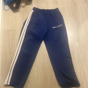 Blå Palm Angels mjukisbyxor med vita ränder - Snygga blå mjukisbyxor från Palm Angels med vita sidoremsor och logotyp på framsidan. Byxorna har elastisk midja, raka ben och dragkedjor vid bensluten. Perfekt för en chill streetwear-look och riktigt sköna att ha på sig.