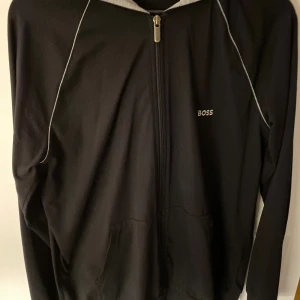 Svart hoodie från BOSS med dragkedja - Svart hoodie från BOSS med grå huva och vita detaljer längs axlarna. Tröjan har hel dragkedja framtill, fickor och BOSS-logga på bröstet. Perfekt för chill dagar och streetwear-stil. Materialet känns mjukt och är troligtvis polyester.