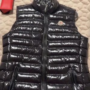 Svart dunväst från Moncler - Snygg svart dunväst från Moncler med glansig finish och quiltad design. Västen har hög krage, två fickor med dragkedja och Moncler-logga på bröstet. Perfekt för lager-på-lager och har en cool, sportig vibe. Dragkedja framtill och klassisk Moncler-etikett på insidan.