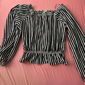 Randig svartvit offshoulder blus - Säljer en svartvit randig blus med offshoulder-modell och långa ärmar. Blusen har resår vid axlar och midja för en snygg passform och lätt volang nertill. Perfekt för dig som gillar trendiga och bekväma plagg med lite edge.