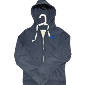 Mörkblå hoodie från Hollister - Mörkblå hoodie från Hollister med dragkedja och vit snörning i huvan. Klassisk logga i blått på bröstet och mjukt material som känns skönt mot huden. Perfekt för chill dagar och enkel att matcha med jeans eller joggers.