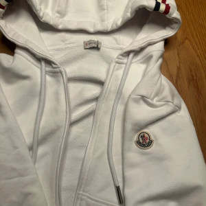 Vit Moncler zip hoodie  - Säljer en clean vit hoodie från Moncler med klassisk logga på ärmen och snygga röd-blå detaljer på huva och muddar. Tröjan har dragkedja, snörning i huvan och är tillverkad i mjuk bomull. Perfekt för dig som vill ha en stilren och exklusiv look.