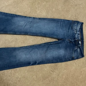 Blå bootcut jeans från Divided, stl 34 - Snygga blå bootcut jeans från Divided i storlek 34. Jeansen har klassisk femficksdesign, coola fickdetaljer med knappar bak och en lätt tvättad look. Perfekta för dig som gillar en avslappnad men trendig stil.