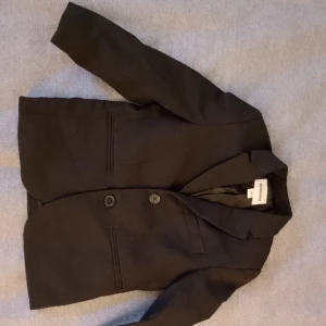 Svart kavaj från H&M - Stilren svart kavaj från H&M med klassisk krage och två knappar framtill. Kavajen har två fickor och är helfodrad, perfekt för en dressad look. Passar dig som vill ha en enkel och snygg kavaj till skolavslutning eller andra tillfällen.