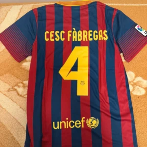 FC Barcelona Fabregas #4 matchtröja Nike - Säljer en officiell FC Barcelona matchtröja från Nike med Cesc Fàbregas och nummer 4 på ryggen. Tröjan är blå och röd med gula detaljer, klassiska vertikala ränder, klubbmärke och Qatar Airways-tryck framtill. Tillverkad i lätt och ventilerande Dri-FIT-material.