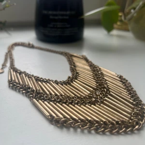 Statement halsband i guldton - Unikt statement halsband i guldton med flera lager av smala metallrör som är sammanlänkade med kedjor. Halsbandet har en bred, V-formad design som ger en cool och edgy look. Perfekt för att lyfta din outfit med en touch av glamour och attityd. #halsband #acessoarer
