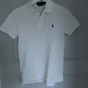 Vit pikétröja från Ralph Lauren - Vit pikétröja från Polo Ralph Lauren i slim fit-modell. Klassisk krage, korta ärmar och två knappar vid halsen. Ikoniska broderade loggan i marinblått på bröstet. Tillverkad i mjuk bomull som är skön mot huden.