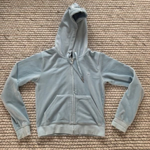 Ljusblå velour hoodie Juicy Couture - Säljer en ljusblå hoodie från Juicy Couture i mjuk velour med dragkedja och huva. Tröjan har långa ärmar, snörning vid huvan och en snygg J-detalj på dragkedjan. Perfekt för dig som gillar klassisk Y2K-stil och vill ha något riktigt comfy.