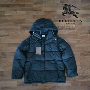 Burberry Puffer - Svart dunjacka från Burberry med klassisk Säljer en elegant och mångsidig Burberry-jacka i svart, i nyskick. Dunfodrad för värme och komfort med det klassiska rutmönstret i huvan som ger en exklusiv touch. Praktiska dragkedjefickor, avtagbar huva och diskret logopatch på ärmen. Passar M/L. Jackan är i princip som ny och utan defekter. En lyxig kombination av stil och funktion – perfekt för både vardag och tillfällen då du vill klä upp dig.  📦Snabb leverans - skickas samma dag📌