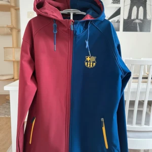 Barça vindjacka i vinrött och blått - Snygg vindjacka från FC Barcelona i tvådelad design med vinröd och mörkblå färg. Jackan har huva, dragkedja, gula detaljer på fickorna och klubbens emblem på bröstet. Materialet är polyester och jackan har normal passform. Perfekt för dig som älskar fotboll och Barça.köpt i Barcelona vid Camp Nou. Helt ny med tagg.