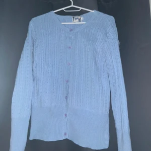 Ljusblå stickad kofta från Joy - Säljer en ljusblå stickad kofta från Joy med kabelstickat mönster och rund hals. Koftan har långa ärmar och matchande blå knappar framtill. Perfekt att slänga över en t-shirt för en chill look. 