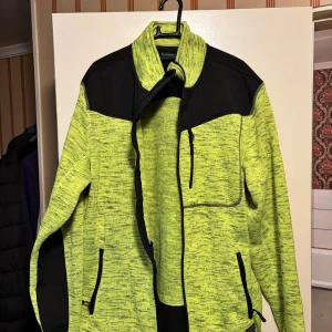 Gul och svart fleecejacka - Säljer en sportig gulmelerad fleecejacka från Blue Wear med svarta detaljer på axlar och fickor. Jackan har dragkedja framtill, tre fickor med dragkedja och hög krage. Perfekt för dig som gillar att sticka ut med färg.