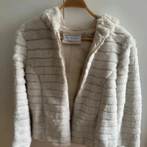 Crèmefärgad fuskpäls jacka - Säljer en fluffig beige pälsjacka med huva och dragkedja framtill. Jackan har horisontella ränder i pälsen och är helfodrad med mjukt tyg på insidan. Perfekt för dig som vill ha en mysig och snygg look under kyliga dagar