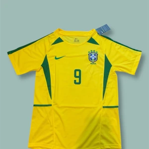 Brasilien Ronaldo 9 fotbollströja - Gul Brasilien fotbollströja med gröna detaljer, nummer 9 och Ronaldo på ryggen. Nike-logga och Brasilien-emblem på bröstet. Tillverkad i lätt, ventilerande polyester med Dri-FIT teknologi för att hålla dig torr. Kortärmad och klassisk passform, perfekt för fans av Ronaldo.