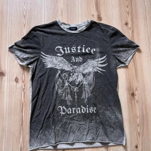 Svart och grå t-shirt från Redwood strl M - Cool t-shirt från Redwood i svart och grått med vit print framtill. Trycket har texten 'Justice and Paradise' och en stor grafisk illustration av en ängel med vingar. Rund hals och korta ärmar, tillverkad i mjuk bomull. Perfekt för dig som gillar edgy streetstyle.