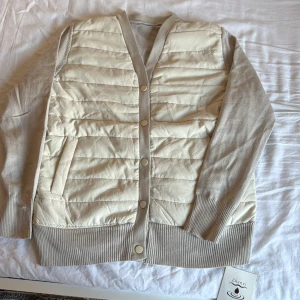 Beige quiltad dunväst från Zhenzi - Quiltad dunväst i beige från Zhenzi med ribbade muddar och knappstängning framtill. Västen har en snygg v-ringad design och två praktiska fickor. Materialmix med dun och stickad polyester ger en cool och mjuk känsla. Perfekt för lager-på-lager under höst och vinter.