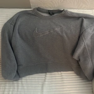 Grå sweatshirt från Nike med logga - Grå sweatshirt från Nike i storlek S med klassisk rund hals och lång ärm. Framsidan har en stor, broderad Nike Swoosh i silvergrått. Tröjan är i mjuk bomullsmix och har en loose passform som är chill och enkel att styla med jeans eller joggers.