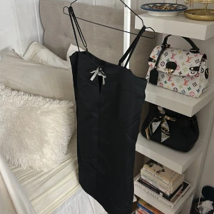 Svart kort klänning med strassrosett - Supersnygg svart kort klänning från H&M med tunna axelband och en glittrig strassrosett framtill. Klänningen har en stilren, figurnära passform och är tillverkad i ett slätt polyestertyg som ger en elegant känsla. Perfekt för dig som gillar att sticka ut med detaljer.