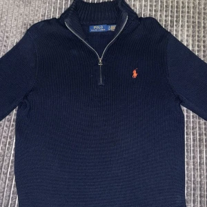 Marinblå stickad tröja Polo Ralph Lauren - Snygg marinblå stickad tröja från Polo Ralph Lauren med half zip och röd broderad logga på bröstet. Tröjan har en ribbad krage och långa ärmar, perfekt för dig som gillar klassisk stil med en modern touch.