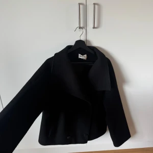 Svart kappa - Säljer en svart kappa från NA-KD med bred krage och dubbelknäppning framtill. Jackan har en kortare, boxig passform och är tillverkad i ett tjockt ullmaterial som håller dig varm under kyliga dagar. Perfekt för dig som gillar klassisk stil med modern twist.