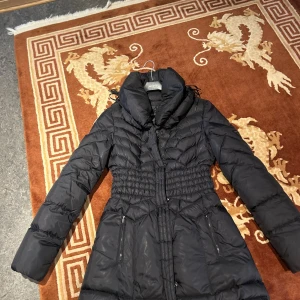 Svart lång dunjacka från Guess - Snygg svart dunjacka från Guess med quiltad design och markerad midja. Jackan har hög krage, dragkedja framtill och två fickor med dragkedja. Materialet är polyester och jackan är långärmad, perfekt för kalla dagar.