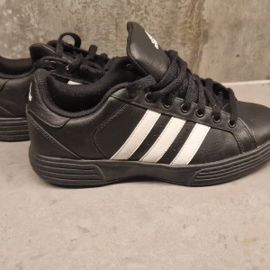 Svarta sneakers från Adidas, strl 37 - Säljer ett par svarta Adidas sneakers med klassiska vita ränder på sidorna. Skorna har rund tå, platt sula och snörning. Ovandelen är i skinn och insidan är fodrad med mjukt textilmaterial. Perfekta för dig som gillar sportig och clean stil.