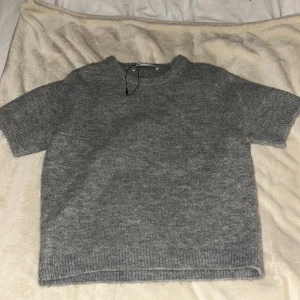 Grå stickad tröja från Zara - Mysig grå stickad tröja från Zara med korta ärmar och rund halsringning. Tröjan har en enkel och stilren design med ribbade muddar vid ärmslut och nederkant. Perfekt för lager-på-lager under hösten eller våren. Passar även till xs
