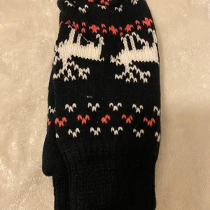 Två par stickade vantar med Fleece Inuti  - Säljer två par stickade vantar. Ena paret är svart med vita och röda detaljer samt renmotiv, perfekta för vintern. Andra paret är lila med vita och rosa hjärtan och mönster, samt snodd runt handleden. Båda paren är varma och mysiga för kalla dagar.  79kr/par 