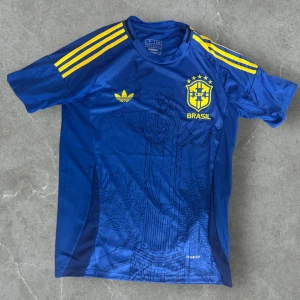 Blå Brasilien fotbollströja Adidas - Snygg blå Brasilien fotbollströja från Adidas med gula detaljer och tre ränder på axlarna. Tryck av Kristusstatyn på framsidan och CBF-logga på bröstet. Tillverkad i lätt och ventilerande material, perfekt för match eller träning.