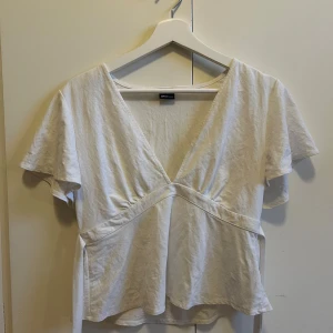 Vit omlottopp med v-ringning och knyt i ryggen💗 - Säljer en vit omlottopp med v-ringning och korta ärmar. Toppen har knytband i midjan som ger en snygg siluett och är tillverkad i mjuk bomull. Perfekt för dig som gillar enkel och clean stil.