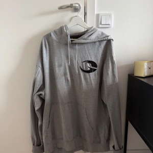 Gasp hoodie - Säljer en grå hoodie med svart broderad logga på bröstet. Tröjan har huva med snörning och en stor magficka framtill. Perfekt för dig som gillar en avslappnad och stilren look.