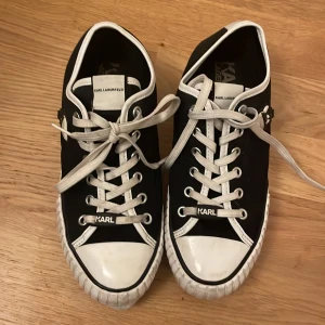 Karl Lagerfeld svarta sneakers - Svarta sneakers från Karl Lagerfeld med vit sula och vita snören. Snygg kontrast med vita detaljer runt tå och kant. På sidan finns en cool Karl Lagerfeld-figur i gummi. Platt sula och rund tå, tillverkade i syntetmaterial. Perfekt för dig som gillar statement-skor med designer-touch.