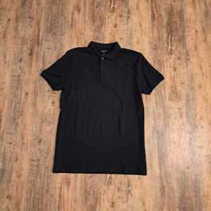 Svart pikétröja från Primark - Klassisk svart pikétröja från Primark med krage och tre knappar framtill. Tröjan är i bomull och har korta ärmar, perfekt för en clean och stilren look. Passar dig som gillar enkel och tidlös stil.