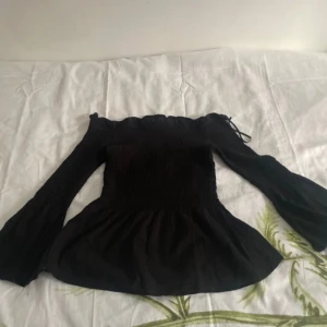 Svart offshoulder blus från Bershka - Supersnygg svart blus från Bershka med offshoulder och långa trumpetärmar. Toppen har smockad överdel och en lätt utsvängd nederkant. Sitter tight upptill Perfekt för dig som gillar trendiga och feminina plagg. Storleken säger M fast den är egentligen mindre💞
