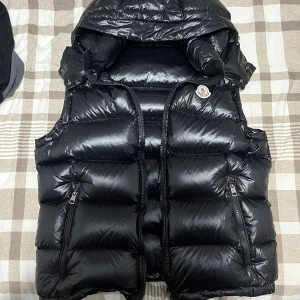 Svart dunväst från Moncler - Snygg svart dunväst från Moncler Storlek S  med glansig finish, hög krage och två dragkedjefickor framtill. Västen har klassisk Moncler-logga på bröstet och coolt serietidningstryck på insidan. Perfekt för lager-på-lager och streetstyle.