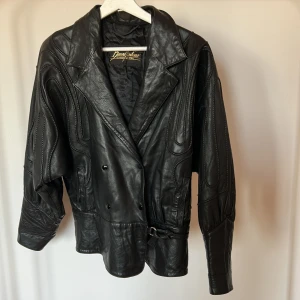Svart skinnjacka från Brant Montana - Unik svart skinnjacka från Brant Montana med coola broderade detaljer på ärmarna, dubbelknäppning och bred krage. Jackan har bälte i midjan och snygga knappar framtill. Står strl 48 men uppfattar mer som en italiensk storlek så 44 är mer sann. Vilket är M-L 