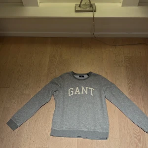 Grå sweatshirt från GANT i S - Snygg grå sweatshirt från GANT med klassisk logotyp i vitt framtill. Tröjan har rund hals, ribbade muddar och långärmad passform. Perfekt för chill dagar och enkel att matcha med jeans eller joggers.