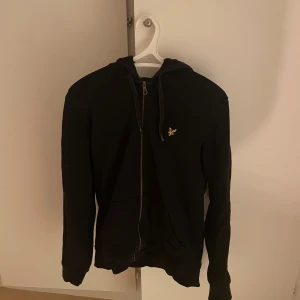 Svart hoodie från Lyle & Scott - Svart hoodie med dragkedja från Lyle & Scott, klassisk gul fågel-logga på bröstet. Tröjan har huva med snörning och två fickor framtill. Perfekt för chill dagar och enkel att matcha med jeans eller joggers.